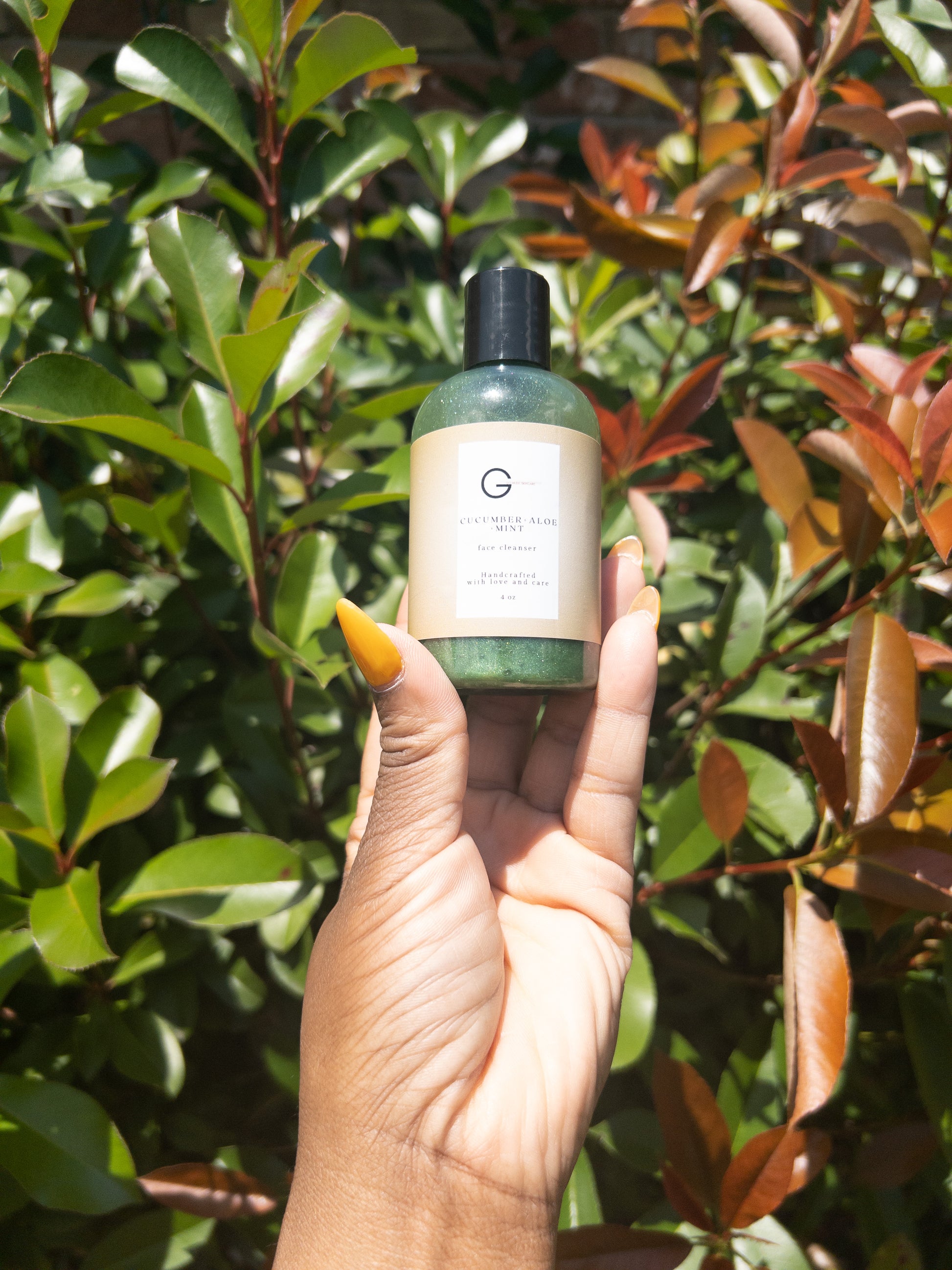 A picture of a woman’s hand holding a 4 oz bottle of cucumber infused face cleanser in front of a bush outside.