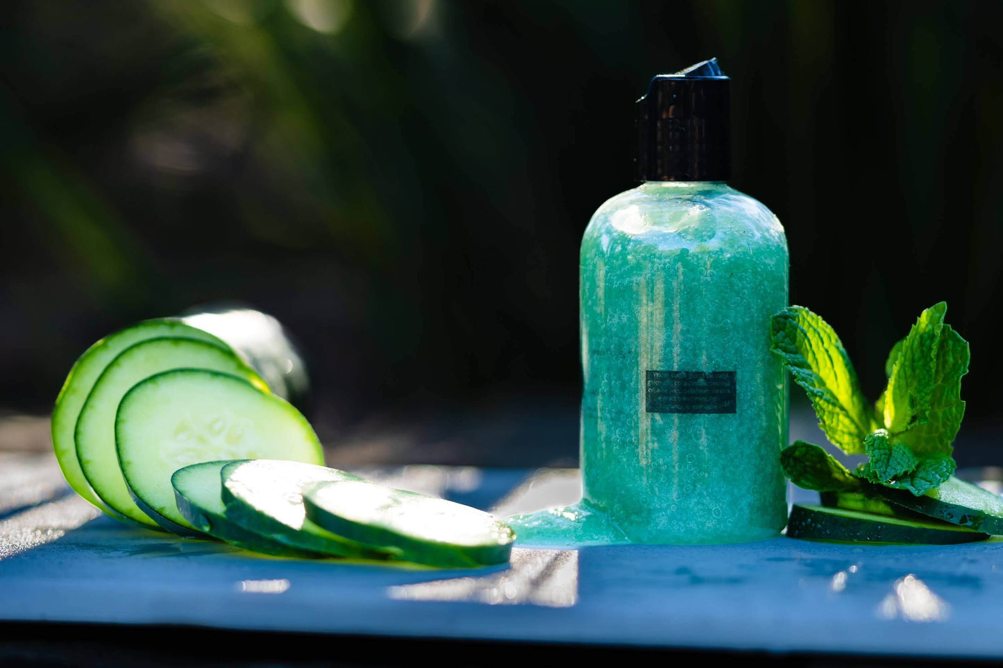 A picture of a 4 oz bottle of green colored cucumber infused face cleanser sitting on a white cutting board with sliced of cucumbers. The sunlight is shining in the background of the picture.