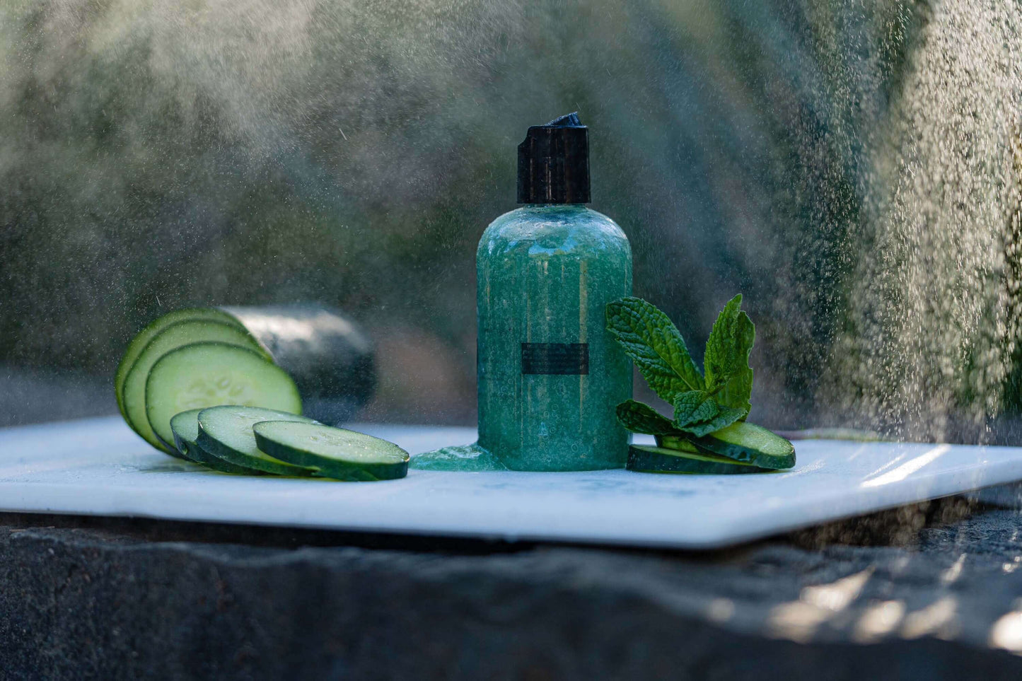 A picture of a 4 oz bottle of green colored cucumber infused face cleanser sitting on a white cutting board with mist in the air.